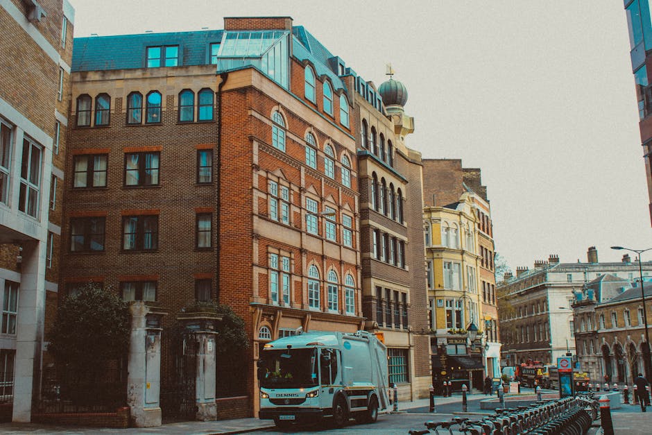 A city street lined with multi-storey buildings displaying a mixture of architectural styles, including red brick facades, ornate window frames, and decorative cornices, set against a light sky. In th