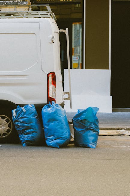 A large pile of black garbage bags, some torn or loosely closed, is stacked against a modern building with a reflective, metallic surface and tiled facade. The bags appear to contain household rubbish